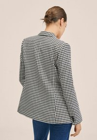 Marynarka w wzór houndstooth w odcieniach czerni i bieli, o zapięciu na jeden guzik, z wyciętymi klapami i guzikiem przy mankietach, wykonana z teksturowanej tkaniny.