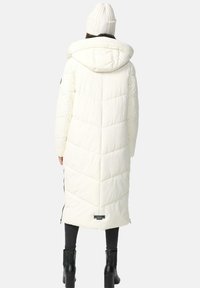 Garšs balts pūkains mētelis ar kapuci, ar ševrona quilted dizainu, sānu rāvējslēdzējiem un logotipu apakšmalā. Saskaņots ar melnajām zābakiem.