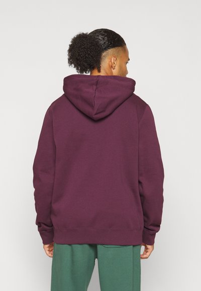 Mörklila hoodie med en stor huva, ribbade ärmslut och rak fåll. Bärs med gröna mjukisbyxor som visar en avslappnad passform och bomullsmaterial.