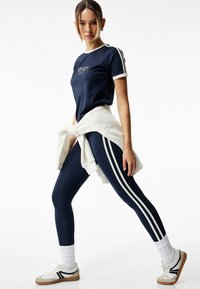 Marineblauw sport T-shirt met witte afwerking en logo, gecombineerd met marineblauwe legging met witte zijkanten. Bevat ook een geknoopte crèmekleurige sweatshirt.