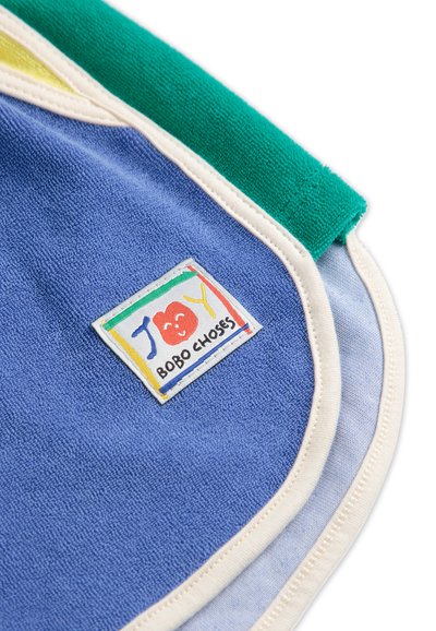 Plava frotirna ručnik s zelenim, žutim i bijelim naglascima; sadrži šarenu oznaku s natpisom "JOY" i "BOBO CHOSES". Mekana, teksturirana površina.