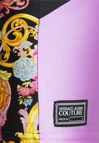Legíny Versace Jeans Couture s živým květinovým vzorem v černé a zlaté na jedné straně a jednobarevným levandulovým provedením na druhé. Připojena černá značka.