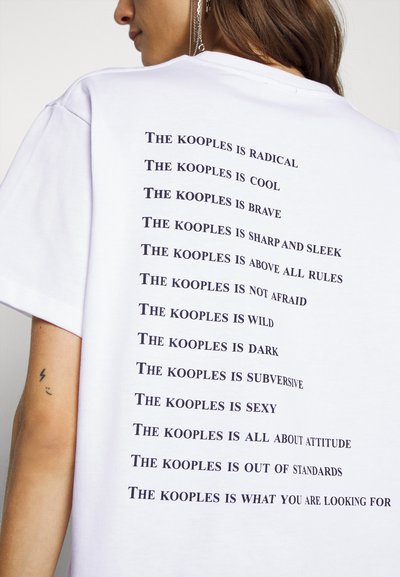 Valge puuvillane t-särk, mille seljal on julge musta teksti trükk, kus erinevad fraasid algavad sõnadega "The kooples is," ja on vertikaalselt kuhjatud.