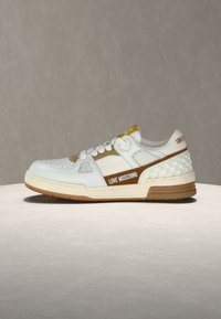 Λευκό και μπεζ αθλητικό παπούτσι Love Moschino με λεπτομέρεια σε σχήμα καρδιάς στα κορδόνια και υφή στη φτέρνα, εμφανίζεται σε προφίλ σε ουδέτερο φόντο.