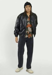 Zwart leren bomberjack met elastische manchetten, gedragen over een oranje en groene camouflage shirt, gecombineerd met donkere jeans en lichte sneakers.