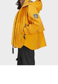 Gelbe wasserdichte Jacke mit Kapuze, versehen mit einem blauen Adidas-Logo auf dem Ärmel und schwarzen Akzenten. Lockere Passform mit verstellbaren Kordelzügen.