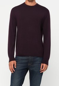 Tummanvioletti neule, jossa pitkät hihat, pyöreä pääntie ja resorit hihansuissa; puettuna sinisten farkkujen päälle. Rinnassa hillitty logo.