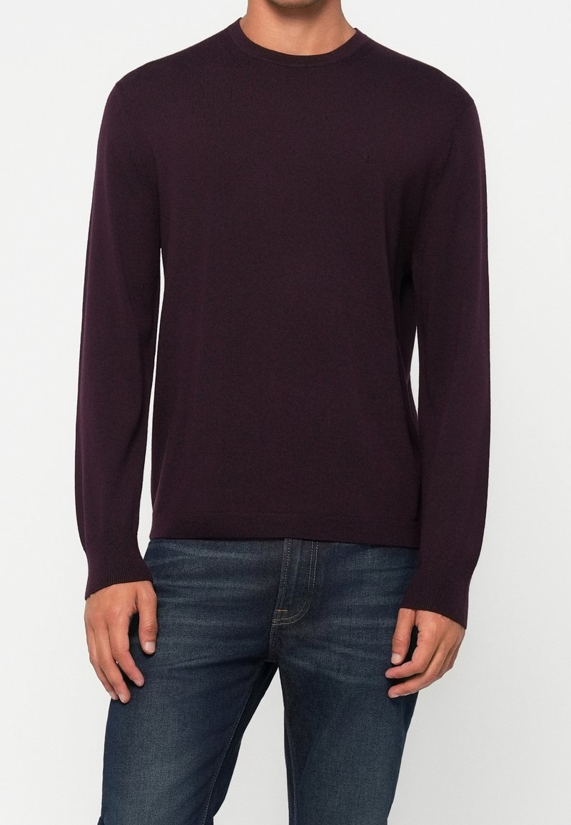 Tummanvioletti neule, jossa pitkät hihat, pyöreä pääntie ja resorit hihansuissa; puettuna sinisten farkkujen päälle. Rinnassa hillitty logo.