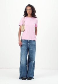 Tricou scurt de culoare roz deschis, cu mânecă scurtă și un logo pe piept, purtat cu blugi largi de culoare albastră și încălțăminte neagră. Modelul ține o geantă mică de culoare bej.