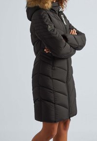 Schwarzer Steppmantel mit gesteppter Musterung, Kapuze mit Kunstfellbesatz und zwei seitlichen Taschen. Verfügt über einen Frontreißverschluss und Logodetails.