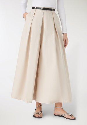 Beżowe spodnie plisowane o szerokich nogawkach z czarnym paskiem, noszone z czarnymi płaskimi sandałami na paski oraz białą długorękawową bluzką, krótką w talii i przy stopach.