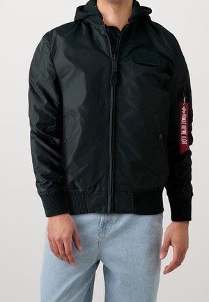 Man draagt een zwarte bomberjack met capuchon en ritssluiting, met geribbelde boorden aan de mouwen en zoom, en een rood 'Remove Before Flight'-label op de zak aan de linkerarm.