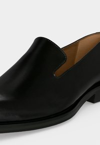 Μαύρο δερμάτινο παπούτσι τύπου slip-on με χαμηλό τακούνι, στρογγυλεμένη μύτη και ταμπά εσωτερική επένδυση, παρουσιασμένο πάνω σε απλό φόντο.