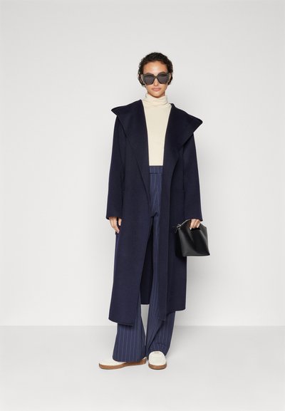 Lungo cappotto blu navy con ampi rever, maglione a collo alto color crema, pantaloni a righe, e scarpe con suola bianca spessa. Borsa nera e occhiali da sole oversize.