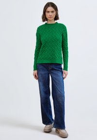 Groene gebreide trui met een gestructureerd zigzagpatroon; gecombineerd met loszittende blauwe spijkerbroek en beige schoenen.