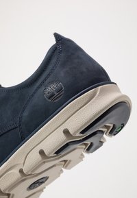 Tumes sinine sametist sneaker, millel on helebeez kummipõhi, tekstuuriga viimistlus ja peenelt väljaulatuv logo kandadel.