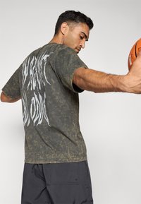 Mand holder en basketball med strakt arm, iført en falmet mørkegrøn T-shirt med stor hvid tekst på ryggen og sorte shorts.