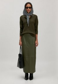 Vrouw met een grijze hoofddoek, zwarte zonnebril, olijfgroene sweater met studs, olijfgroene lange rok, zwarte laarzen, die een zwarte handtas vasthoudt tegen een lichte achtergrond.