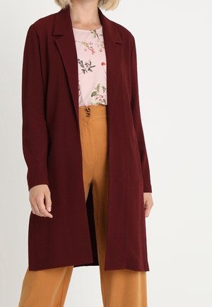 Kvinde iført en lang burgunder åben cardigan over en blomsterprintet lyserød bluse og sennepsgule bukser med højt talje.
