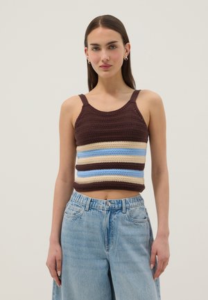 Jeune femme portant un débardeur tricoté rayé marron, bleu et beige et un jean taille haute bleu clair, debout devant un fond uni.