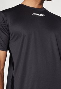 Czarna sportowa koszulka wykonana z lekkiego materiału. Posiada okrągły dekolt, krótkie rękawy oraz biały napis "HUMMEL" na piersi.