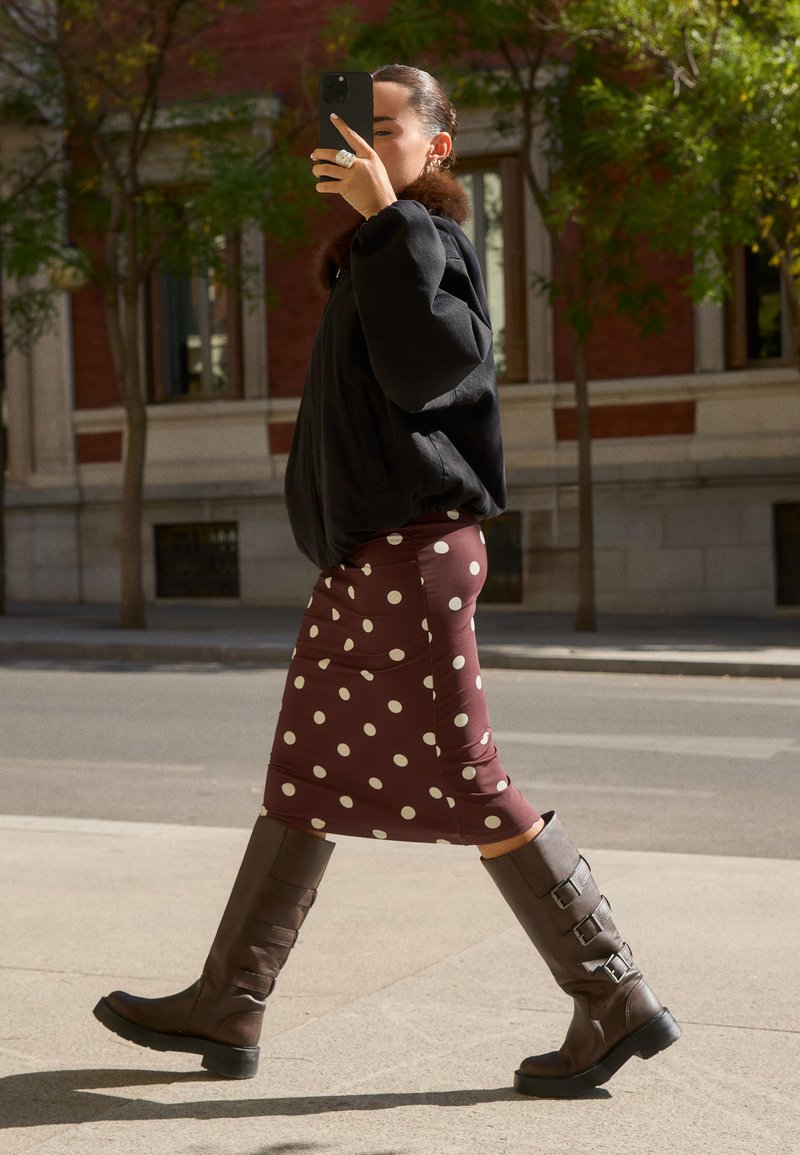 Veste bomber noire, jupe midi à pois bordeaux, et bottes marron jusqu'aux genoux avec boucles. Pose en marche sur un trottoir avec de la verdure en arrière-plan.