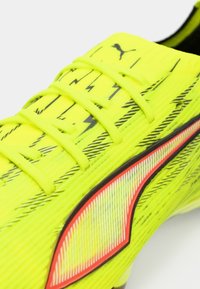 Zapatilla deportiva amarilla con parte superior texturizada con patrones de rayos negros, una franja de acento roja y cierre tradicional de cordones.