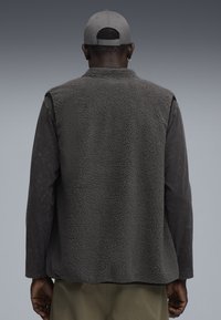 Grau gestrickte Fleeceweste mit strukturierter Oberfläche, getragen über einem dunklen Langarmshirt. Der Rücken hat ein einfaches Design ohne Verzierungen.