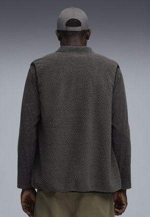 Grau gestrickte Fleeceweste mit strukturierter Oberfläche, getragen über einem dunklen Langarmshirt. Der Rücken hat ein einfaches Design ohne Verzierungen.