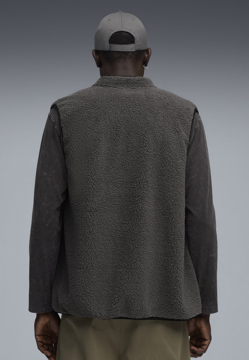 Grau gestrickte Fleeceweste mit strukturierter Oberfläche, getragen über einem dunklen Langarmshirt. Der Rücken hat ein einfaches Design ohne Verzierungen.