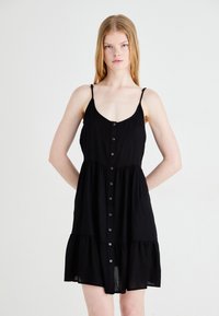 Rochie neagră fără mâneci, cu nasturi, cu un trunchi ajustat, fustă în straturi și bretele ajustabile; realizată dintr-un material ușor, texturat.