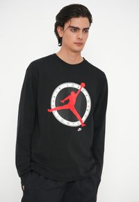 Μαύρο κοντομάνικο πουκάμισο με μακριά μανίκια που διαθέτει μια μεγάλη γραφική παραστάσεις Air Jordan σε κόκκινο και λευκό χρώμα. Μαλακό ύφασμα και άνετη εφαρμογή με στρογγυλή λαιμόκοψη.