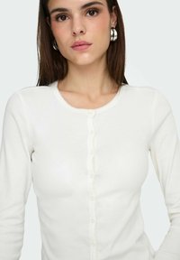 Haut blanc à manches longues côtelées avec un col rond, une bordure texturée et une patte de boutonnage à six boutons avec des bords festonnés. Tissu lisse.