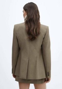 Blazer à carreaux marron avec un design structuré, des revers crantés et une fente simple à l'arrière. La texture semble lisse avec des accents de boutons.