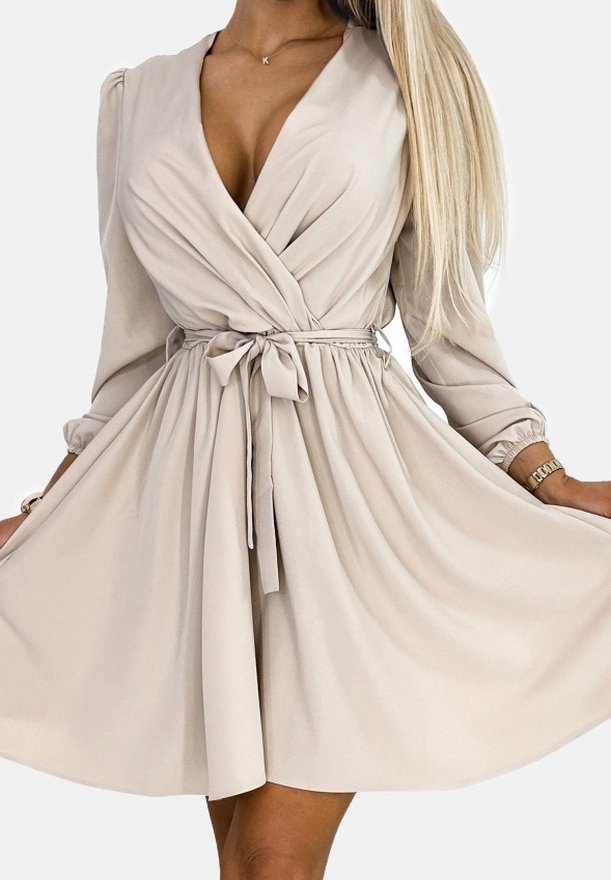 Femme portant une robe portefeuille beige à manches longues avec un décolleté en V et une ceinture nouée à la taille, tenant la jupe écartée sur les côtés.