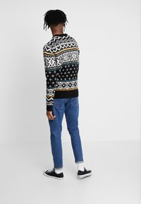 Pull en tricot à motifs noir, blanc, avec des accents de turquoise et d'or, associé à un jean bleu et des baskets noires. Vue de l'arrière.