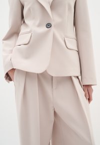 Beige blazer s jedním černým knoflíkem, dvěma předními kapsami a ostrými klopami, spárovaný s odpovídajícími skládacími kalhotami. Hladká, dobře padnoucí tkanina.
