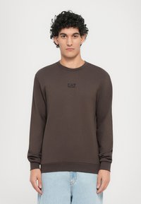 Felpa marrone a maniche lunghe con polsini e orlo a coste, con un piccolo logo nero sul petto; abbinata a jeans azzurri chiari.