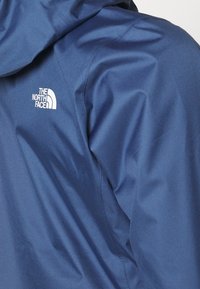 Gros plan d'une veste d'extérieur bleue avec capuche montrant un logo blanc The North Face sur la partie supérieure gauche de la poitrine.