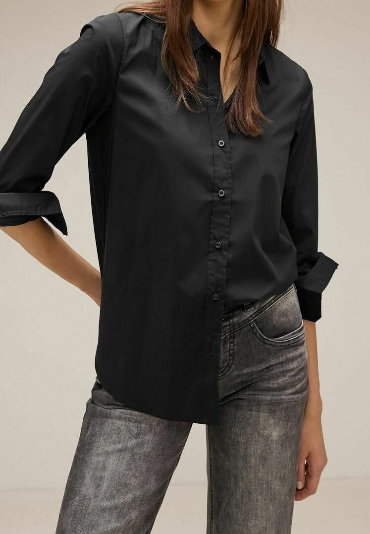 Femme portant une chemise noire à boutons avec les manches retroussées et un jean gris délavé sur un fond clair uni.