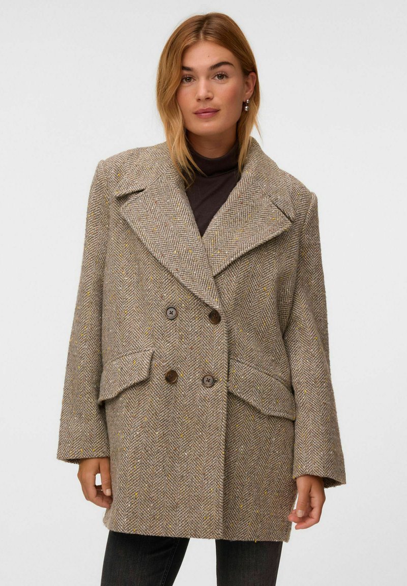 Femme portant un manteau en laine marron à chevrons croisé avec de larges revers par-dessus un haut noir sur un fond uni.