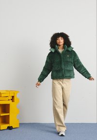 Groene corduroy pufferjas met een hoge kraag en drukknoopsluitingen, gedragen over een beige top en bijpassende lichte broek. Schoenen met rubberen zolen.