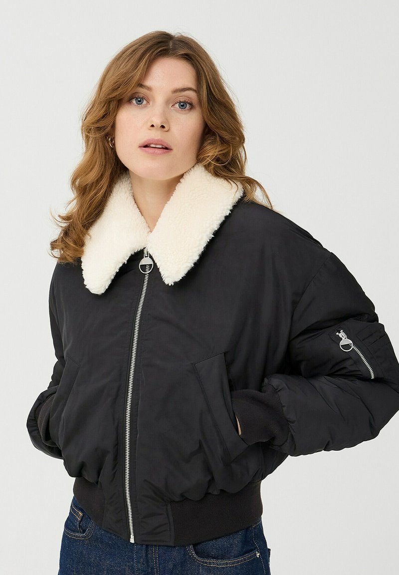 Czarna kurtka bombowca z kołnierzem z kremowego sztucznego owczego futra, zapinana na zamek, z ściągaczami przy mankietach i bocznymi kieszeniami. Posiada małą kieszeń z zamkiem błyskawicznym na rękawie.
