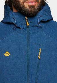 Niebieska kurtka odporna na warunki atmosferyczne z kapturem, z zamkiem błyskawicznym z przodu, pomarańczowymi detalami i skośną boczną kieszenią. Logo wyeksponowane na piersi.