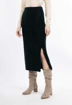 Frau trägt einen schwarzen, gestrickten Midirock mit seitlichem Schlitz, beiges Langarmoberteil und beige Wildleder-Stiefel mit mittelhohem Absatz.