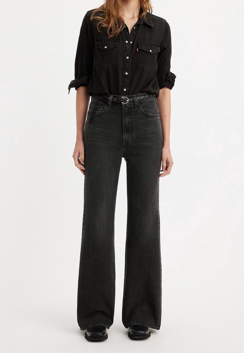 Femme portant une chemise noire à boutons, un jean noir taille haute à jambes larges, une ceinture tressée et des chaussures noires sur un fond blanc.