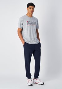 Camiseta gris de manga corta con "Champion Athletic Wear" impresa en negro y rojo. Pantalones deportivos azul marino, zapatillas blancas con detalles rojos.