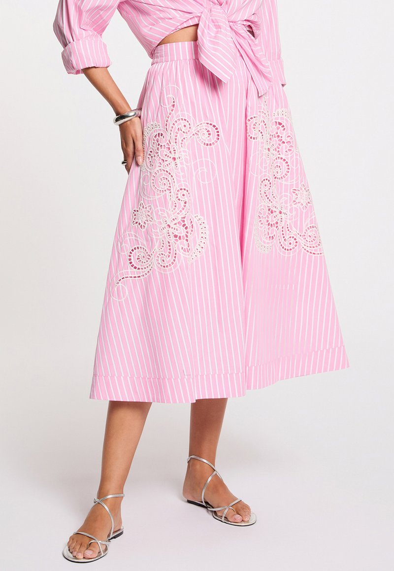 Mujer que lleva una falda midi de rayas rosas y blancas con bordado floral blanco, combinada con sandalias plateadas de tiras y una blusa rosa a juego con nudo.