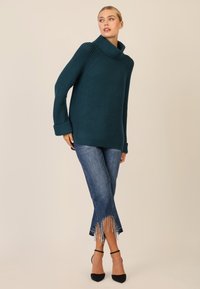 Maglione in maglia teal con collo alto a tubo e maniche lunghe, abbinato a jeans con orlo sfilacciato decorati con dettagli a perle. Tacchi neri.