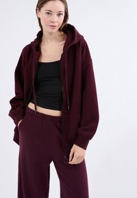 Felpa con zip color burgundy, dotata di un ampio cappuccio, morbida al tatto, dalla vestibilità morbida e con polsini elasticizzati, abbinata a pantaloni comfy di taglio rilassato.